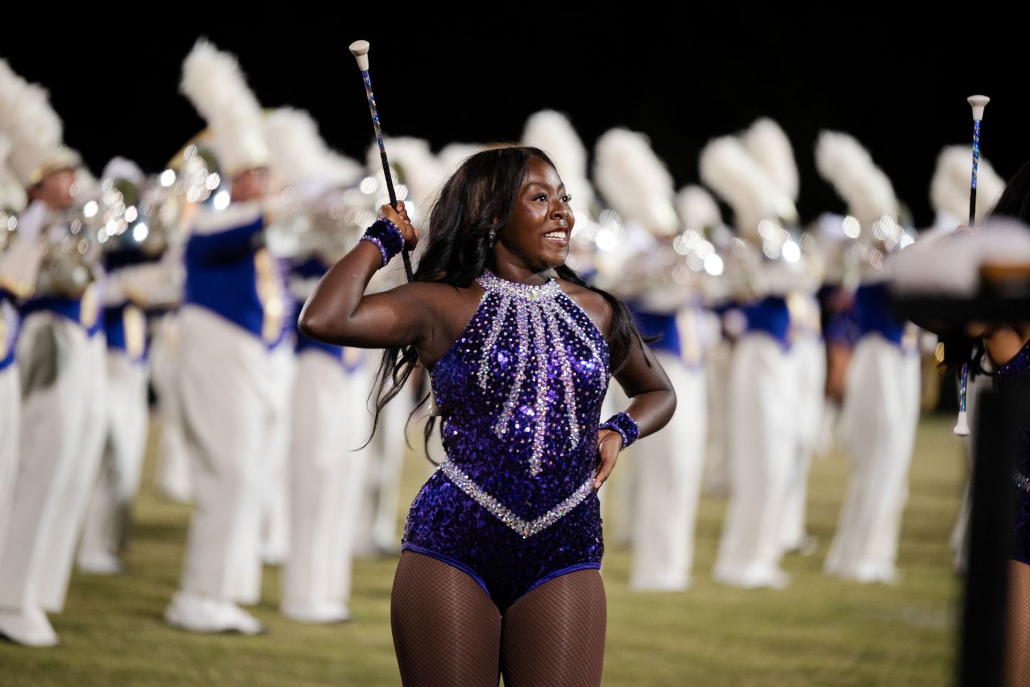 A baton twirler in a sparkling purple leotard performs in front of a marching band at night.