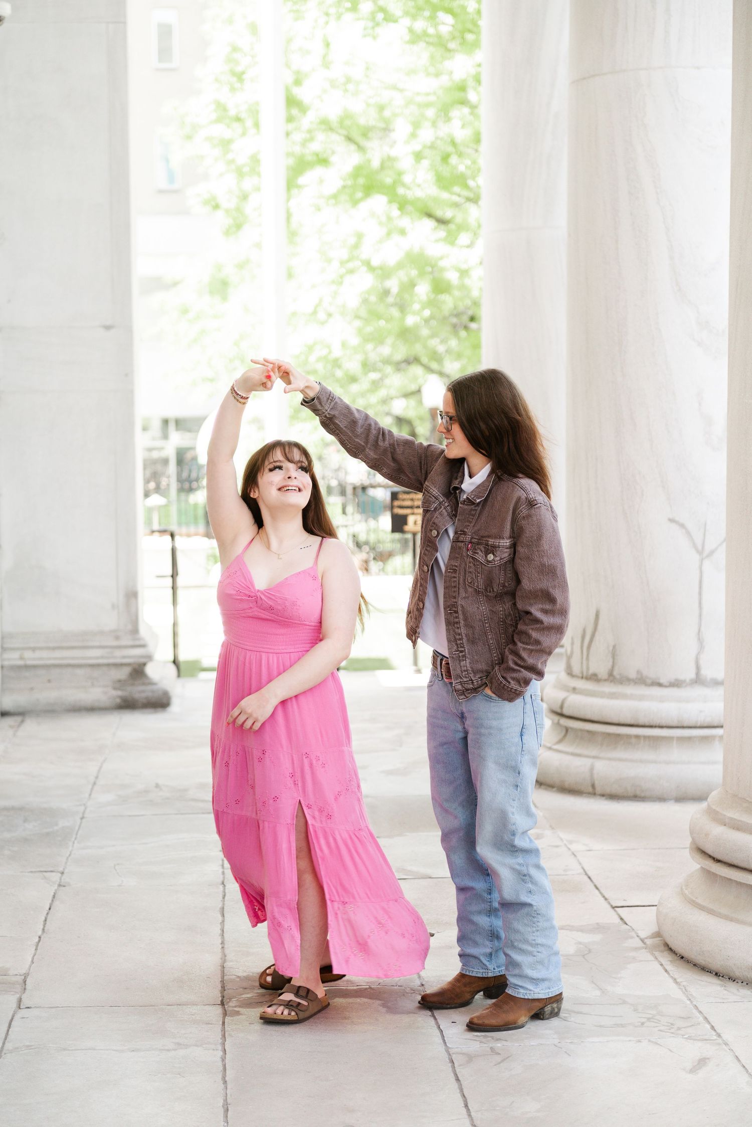 Couple dancing outdoors near white columns, woman in pink maxi dress being twirled by partner in denim jacket.