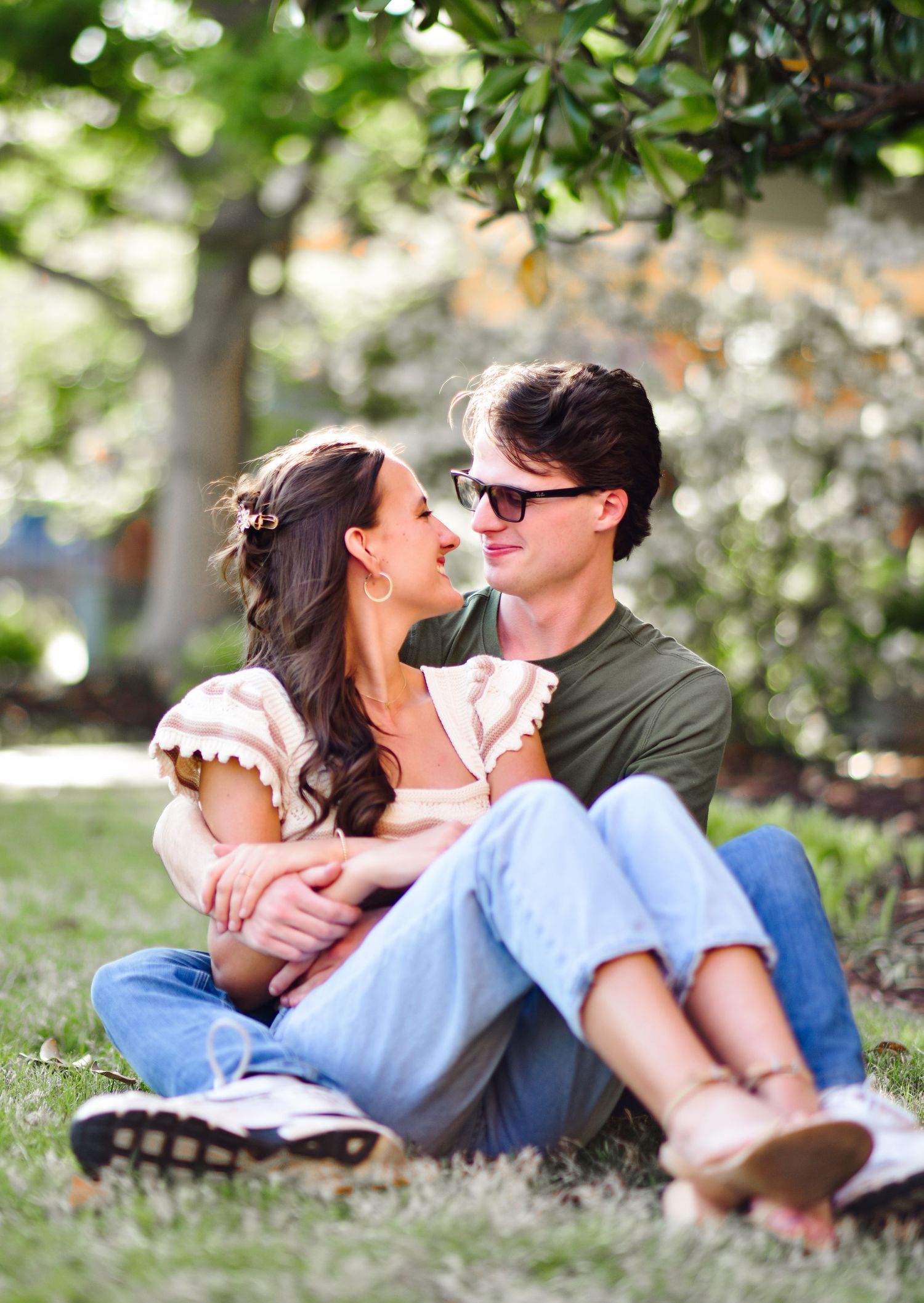 Young couple sitting on grass under a tree, gazing at each other romantically during an outdoor engagement photo session.