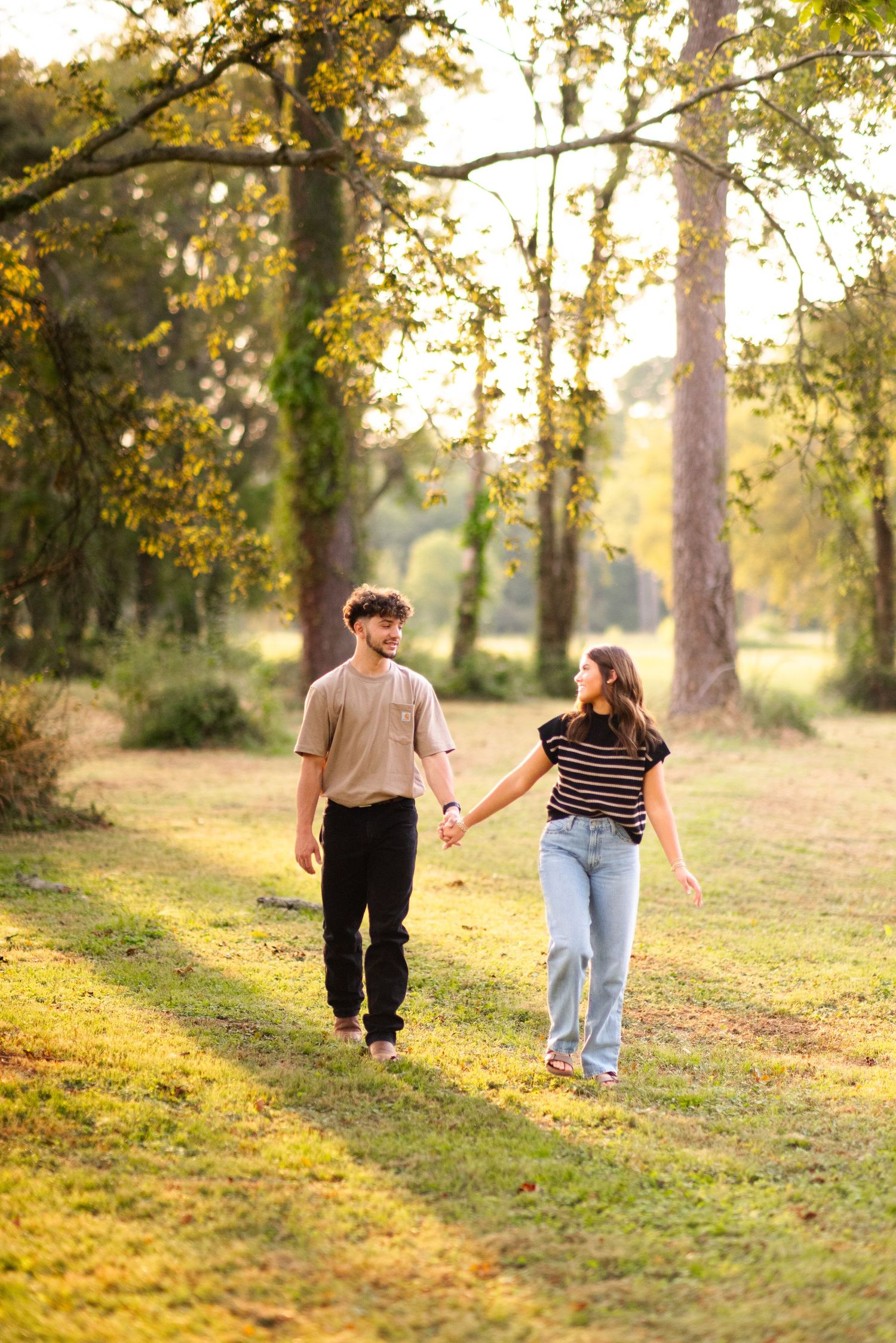 Young couple holding hands and walking through a sunny park with tall trees in background.