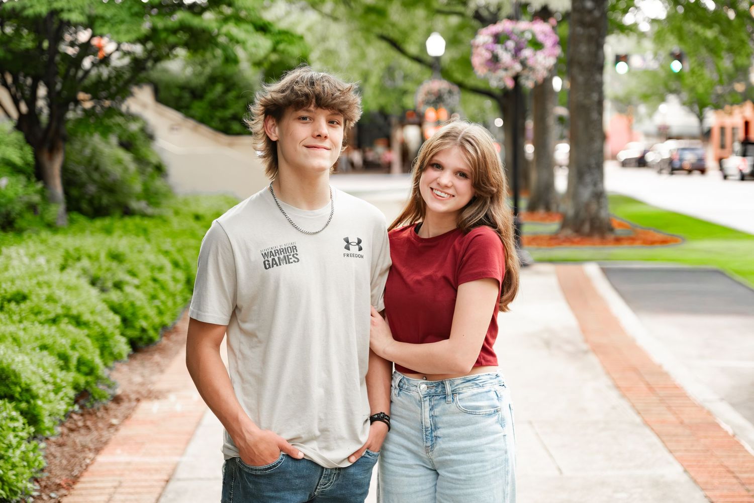 Teen boy and girl in casual outfits smiling together on a brick-lined outdoor walkway with trees.