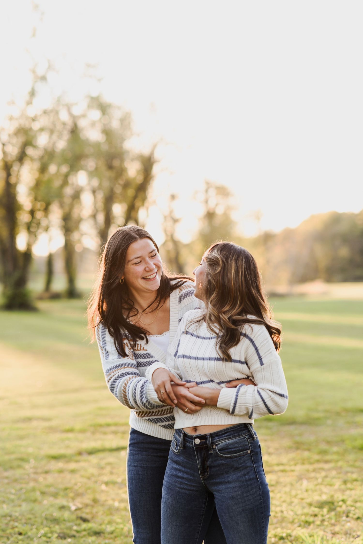 Two women laughing and hugging in a sunlit green park during outdoor photo session.