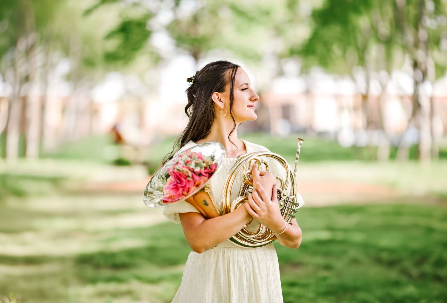 Young woman in floral white dress holding French horn and flowers smiling in green park.