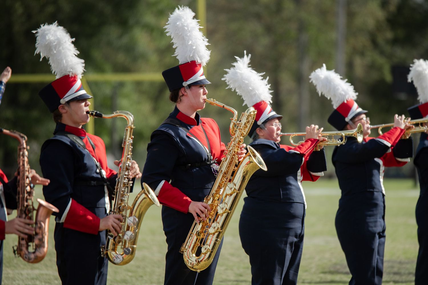 Marching band saxophonists in red and black uniforms with white feathered hats performing outdoors.