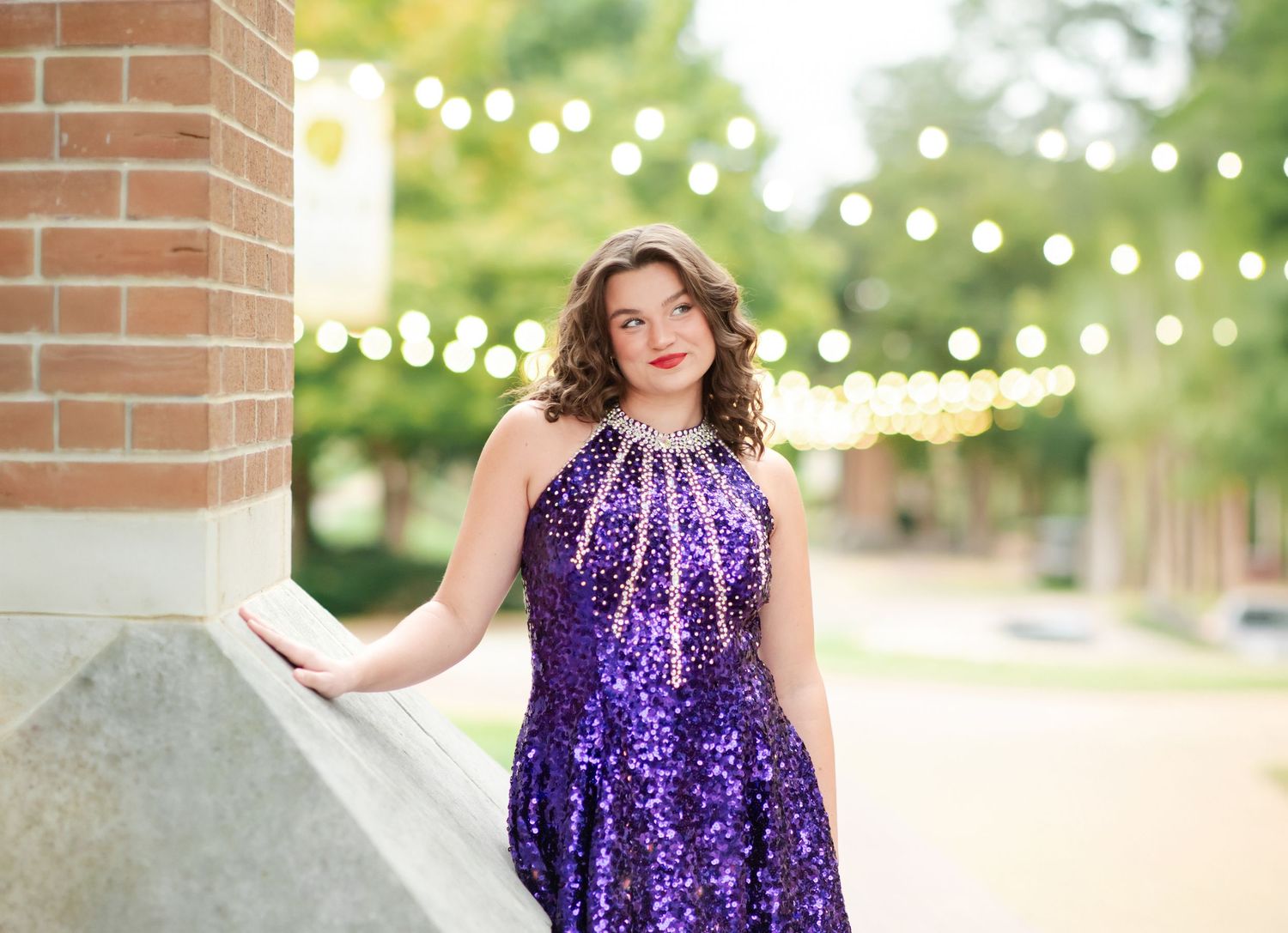 Young woman in a sparkling purple sequined dress leaning against a brick wall with bokeh string lights.