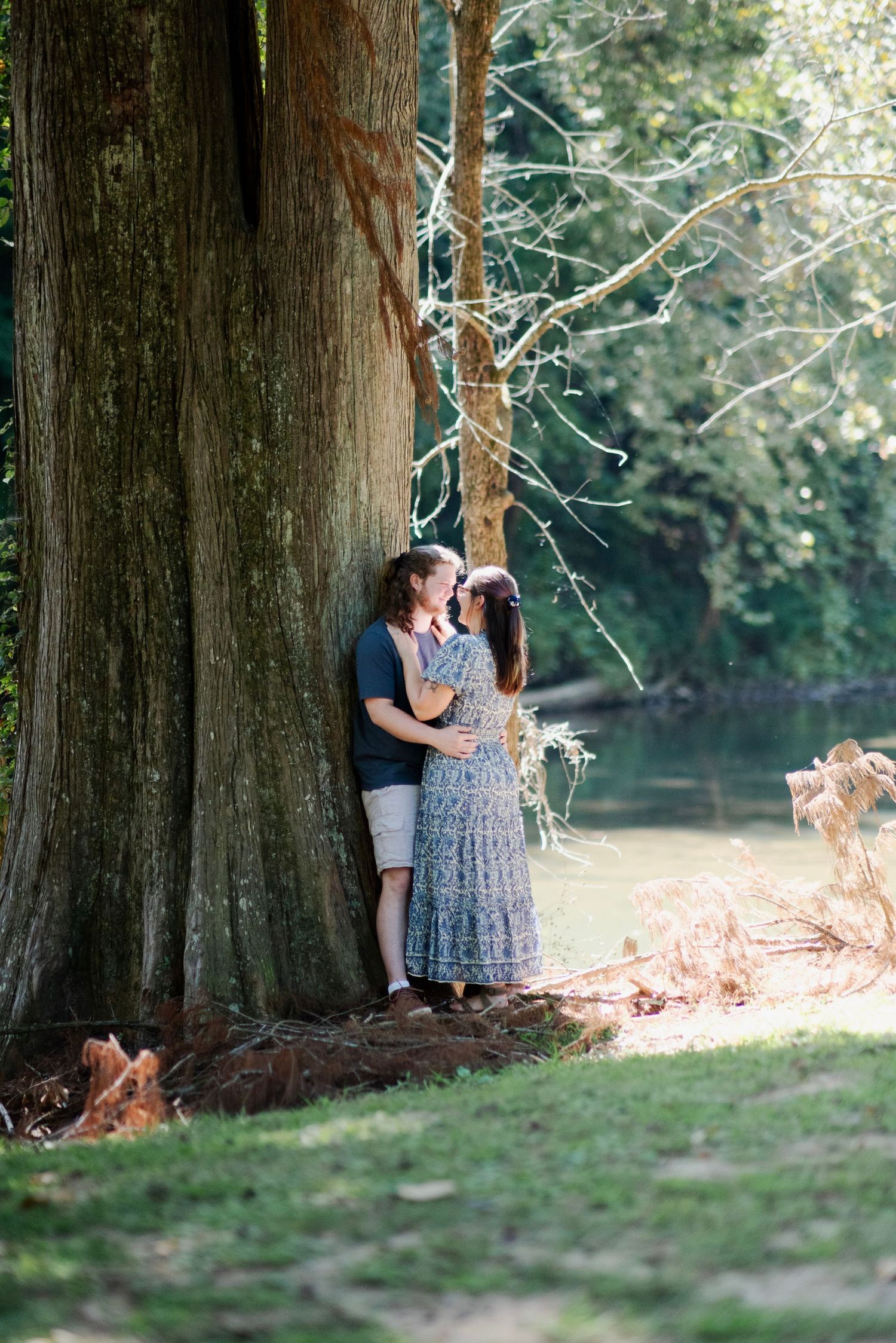 Romantic couple standing close together leaning against a large tree near a peaceful river.