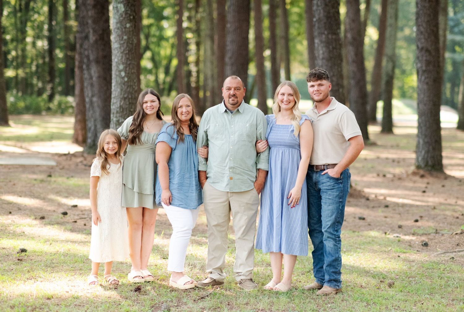 A family of six poses together smiling in a pine forest wearing coordinated earth-tone outfits.