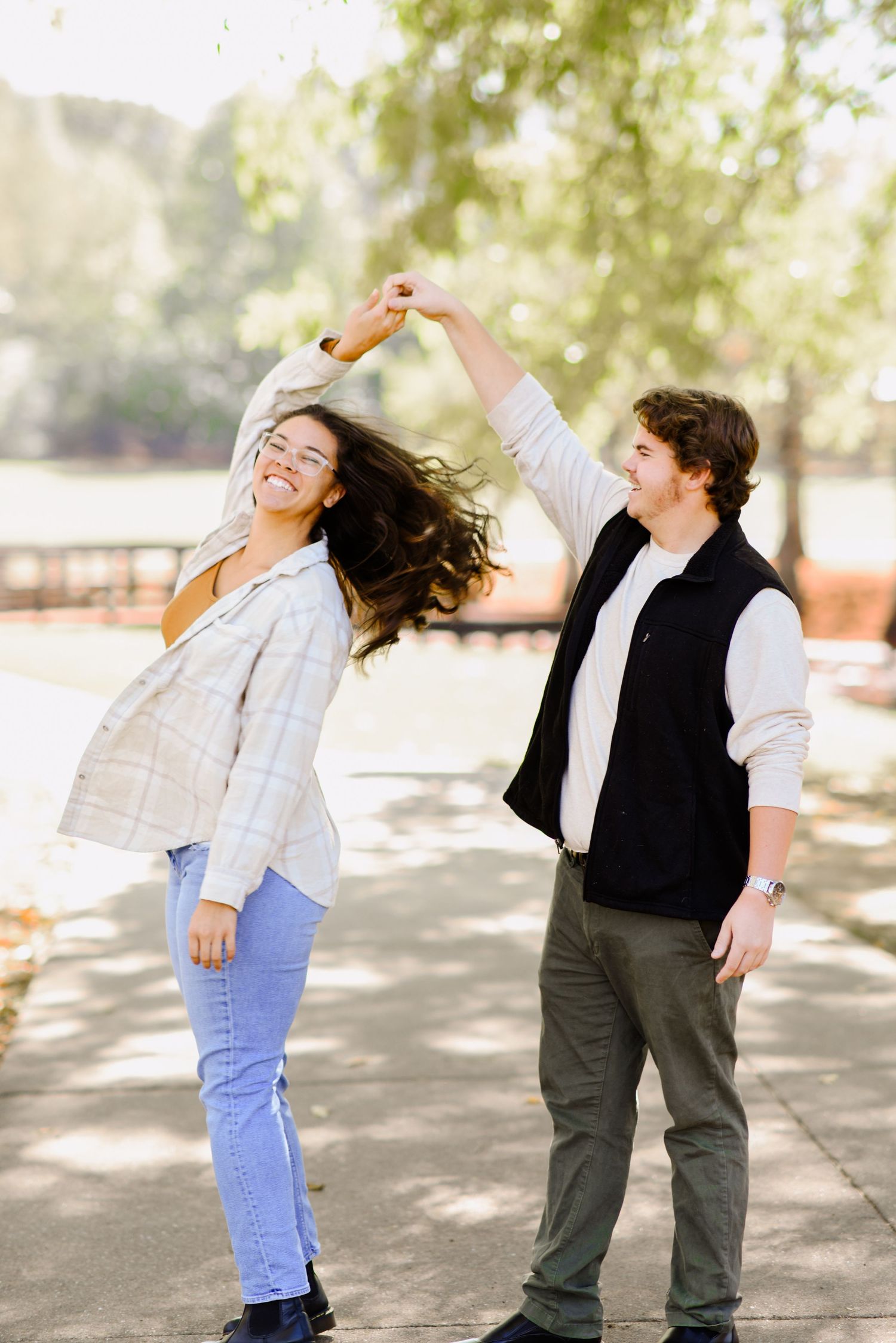 Joyful couple dancing outdoors in a park, man twirling woman with long flowing hair, both laughing in casual fall outfits.