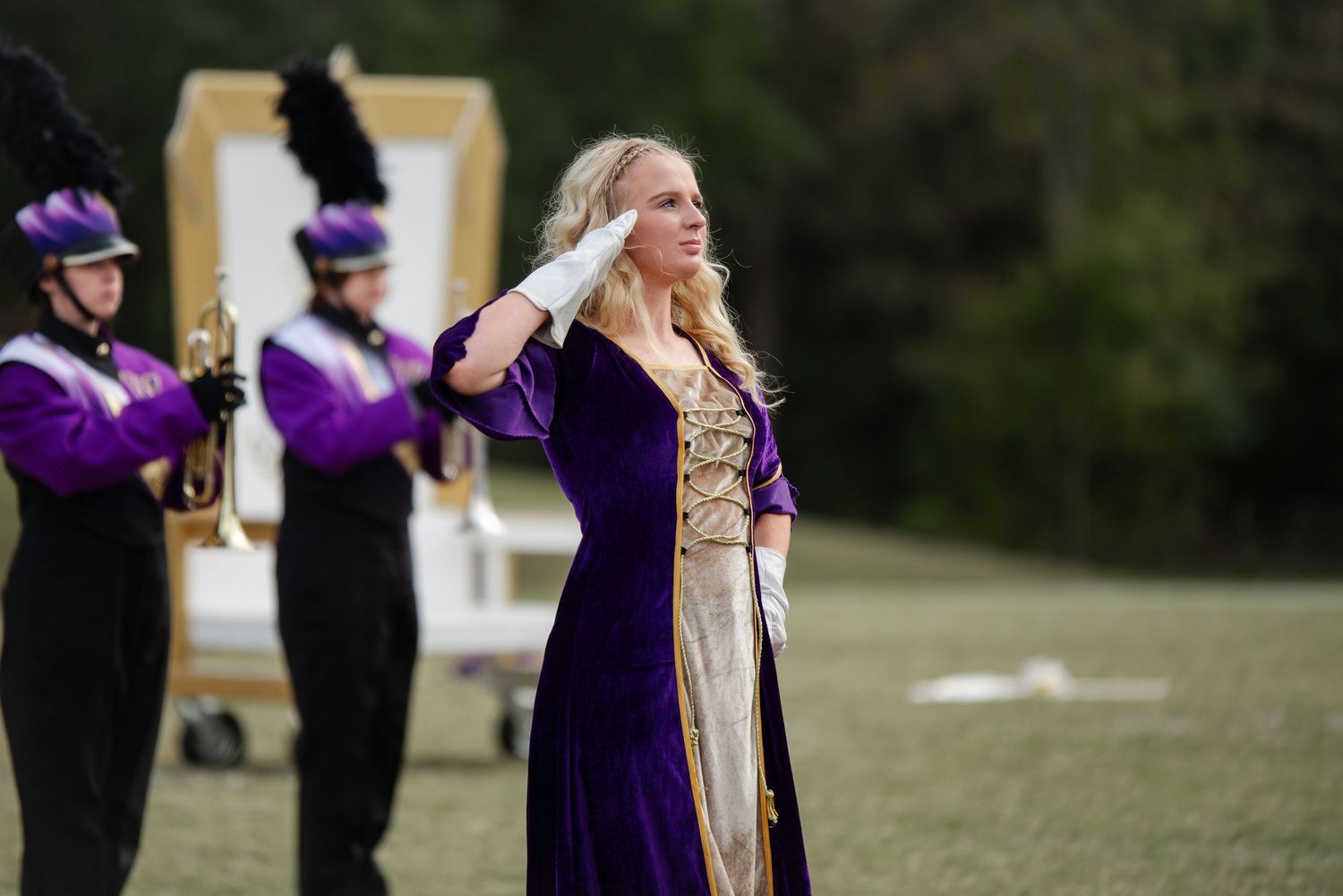 Marching band drum major in purple velvet gown saluting during outdoor field performance.