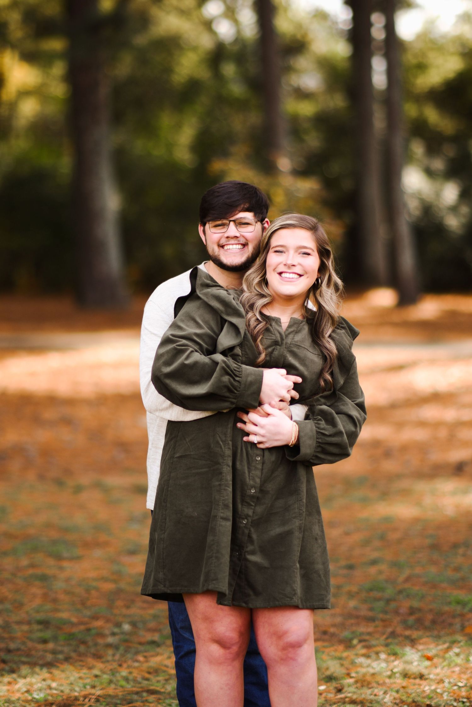 Happy couple hugging in a pine forest, man in white shirt embracing woman in olive green dress, both smiling warmly.