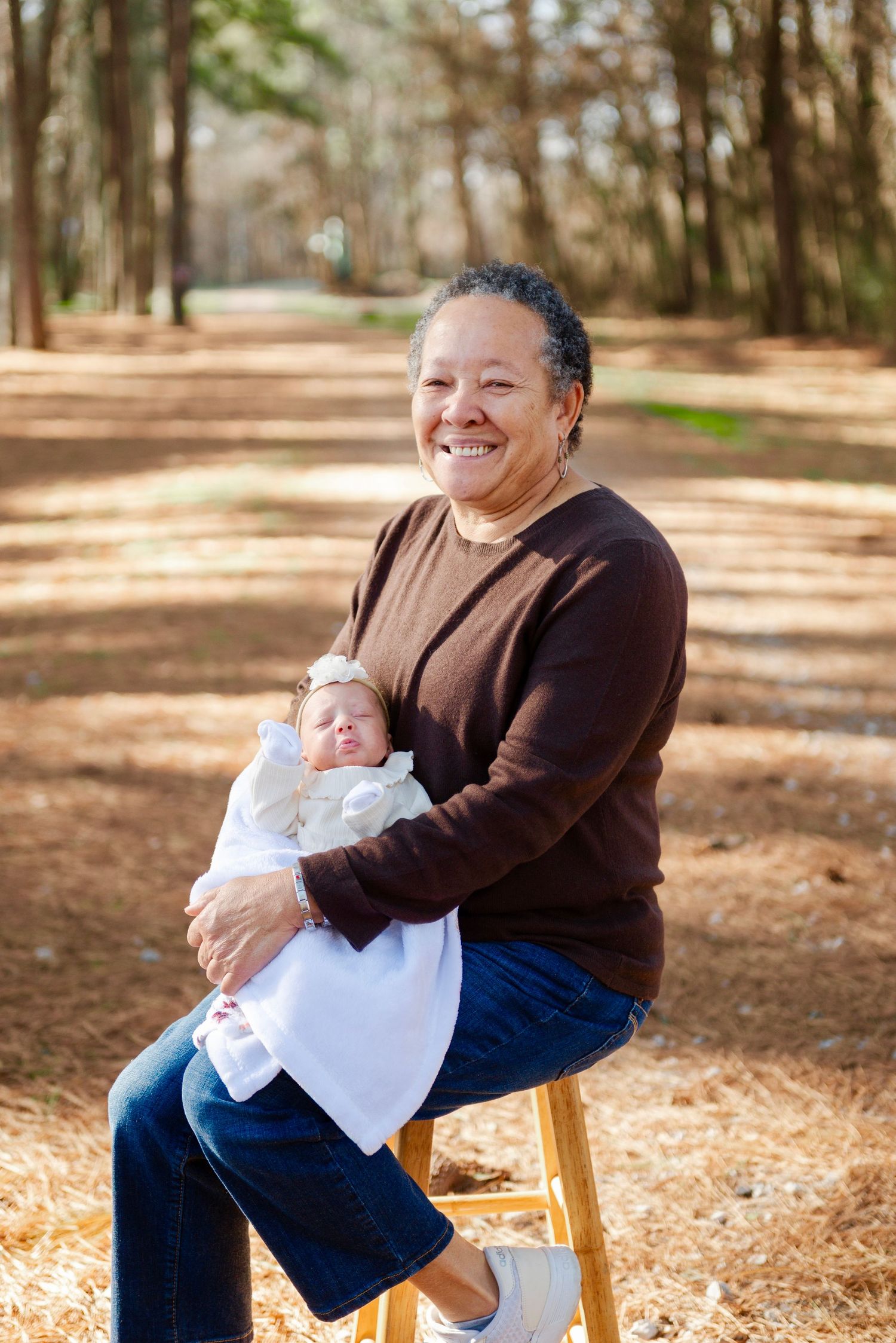 Smiling grandmother sits on stool holding sleeping newborn baby dressed in white outdoors.