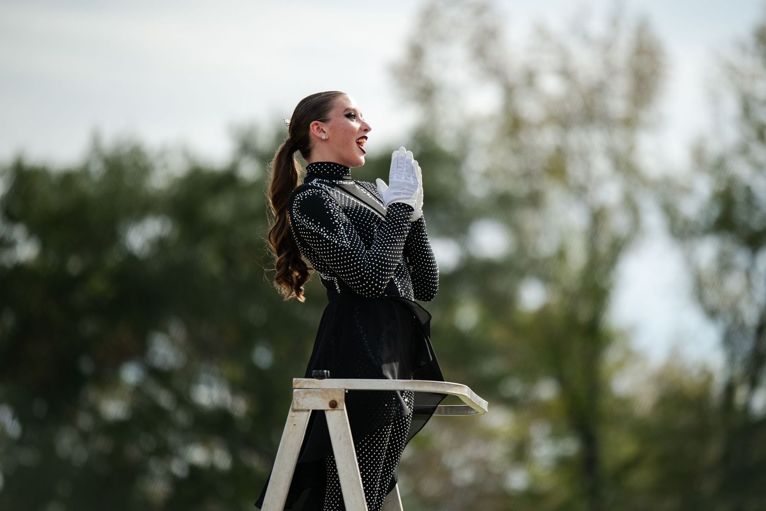 Female band conductor in black uniform stands on ladder during outdoor performance.