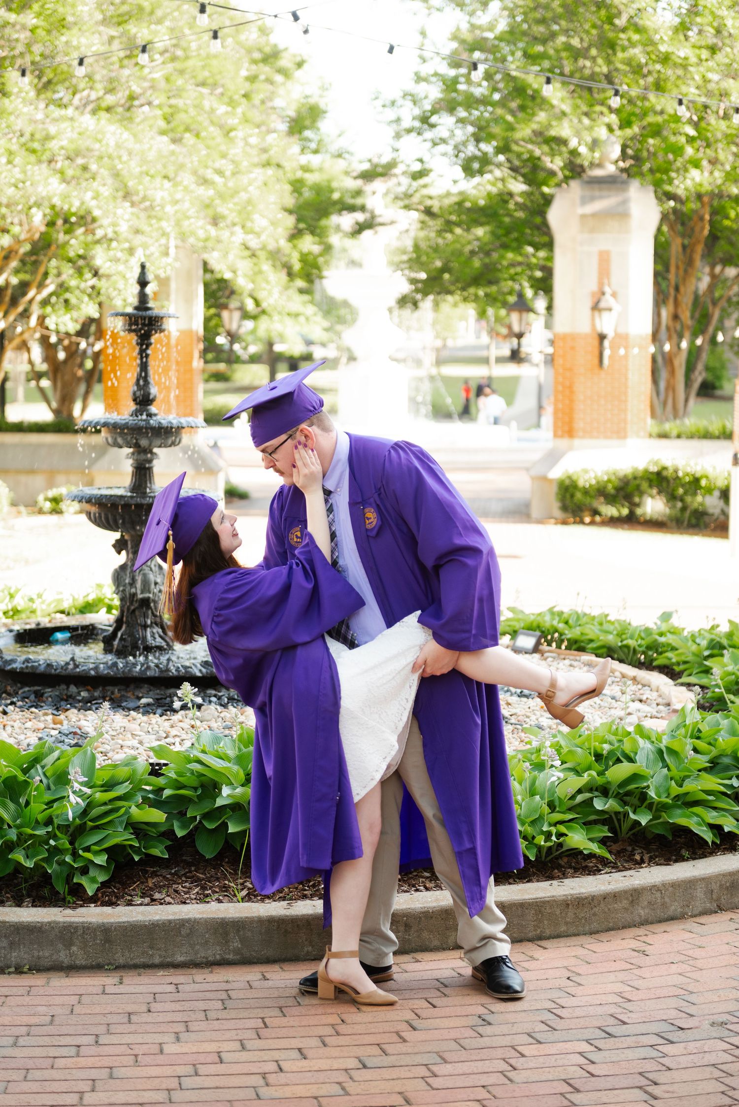 Couple in matching purple graduation gowns share romantic dip near campus fountain garden.