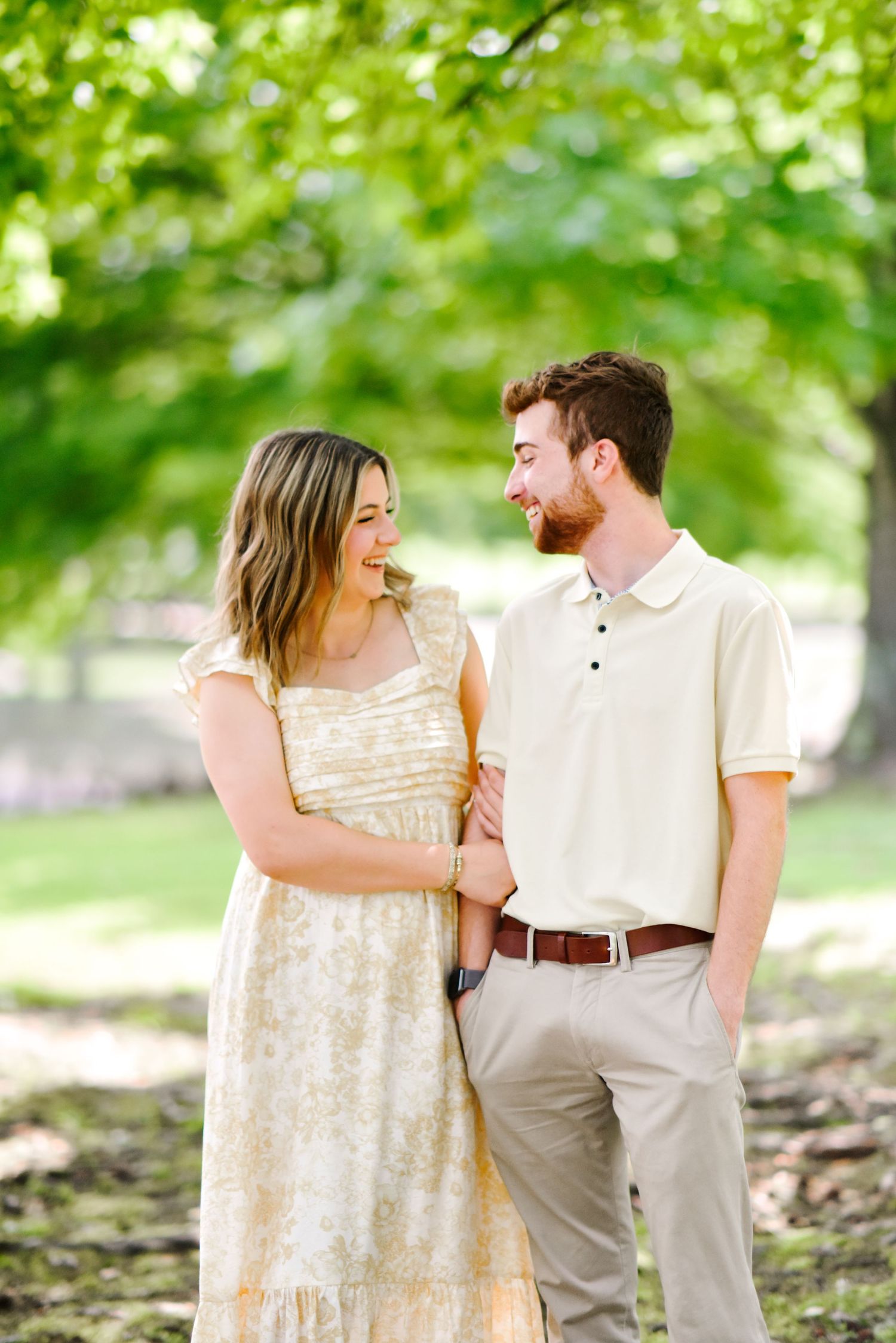 Laughing couple standing together under green trees, woman in yellow floral dress holding mans arm, man in cream polo shirt.