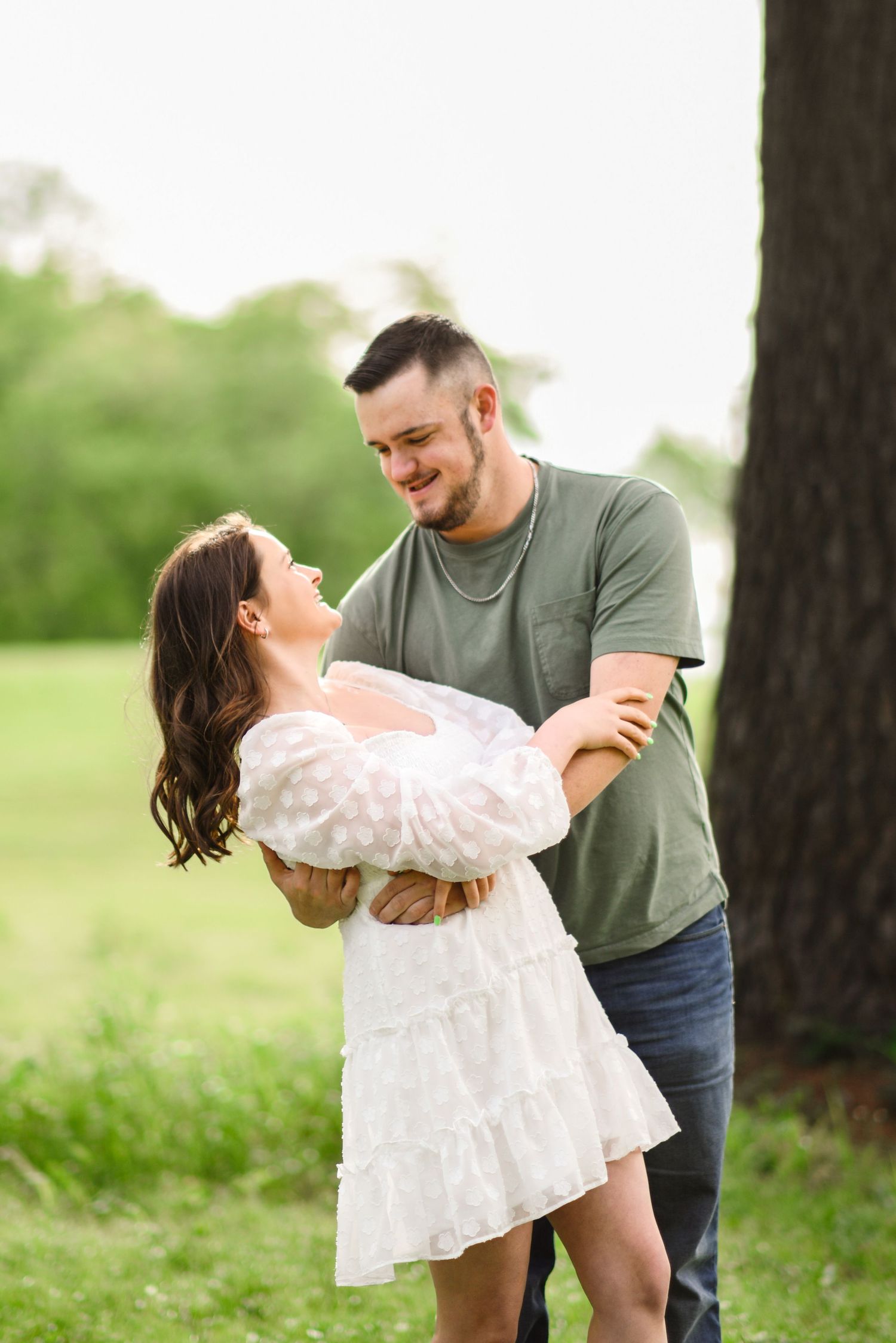 Happy couple dancing near a tree in a green field, woman in white dress, man in olive shirt and jeans, smiling joyfully.