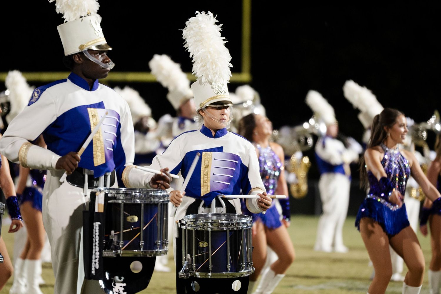 Marching band drummers in blue and white uniforms performing at night with color guard dancers.