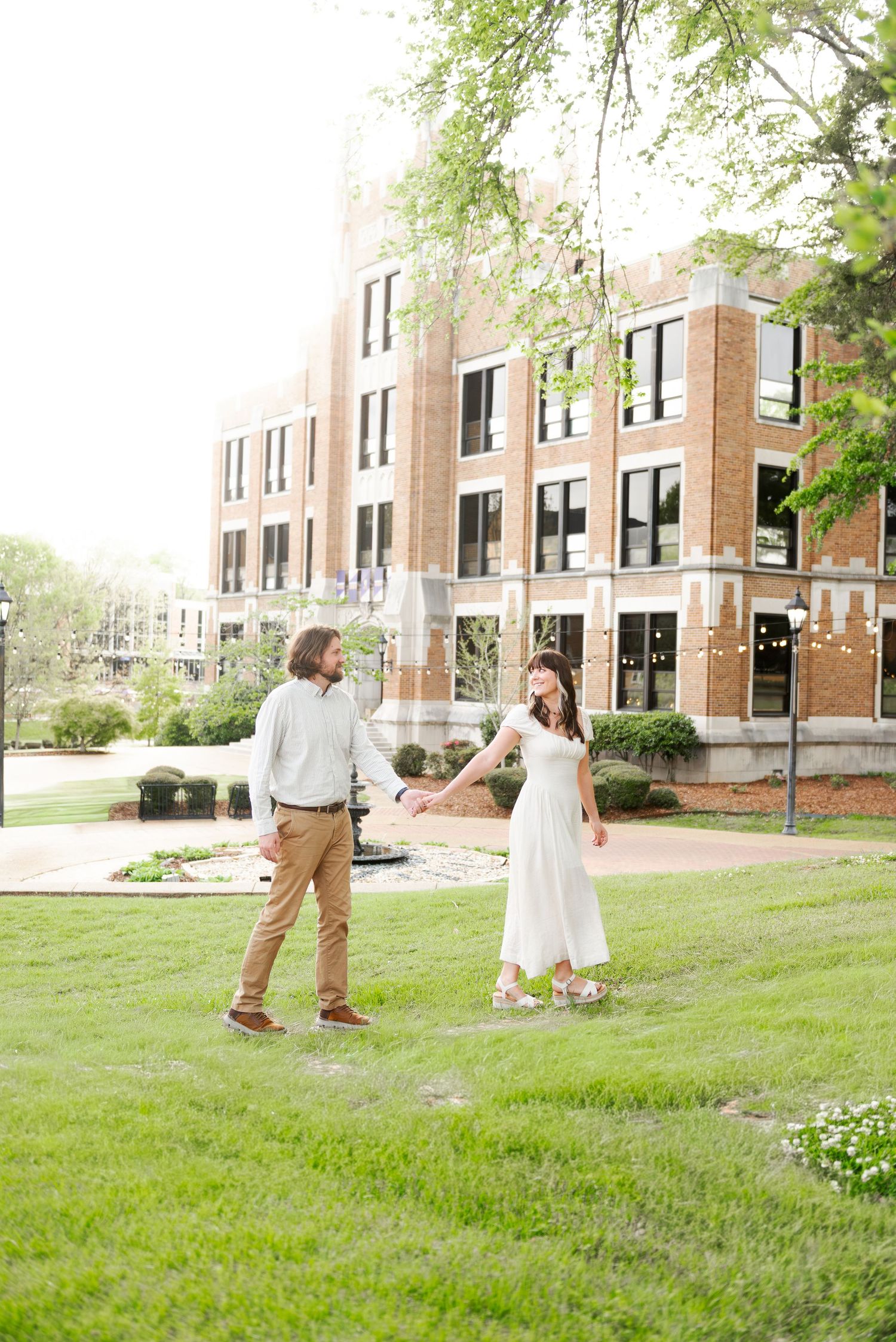 A couple holds hands and walks across a green lawn outside a large brick university building.