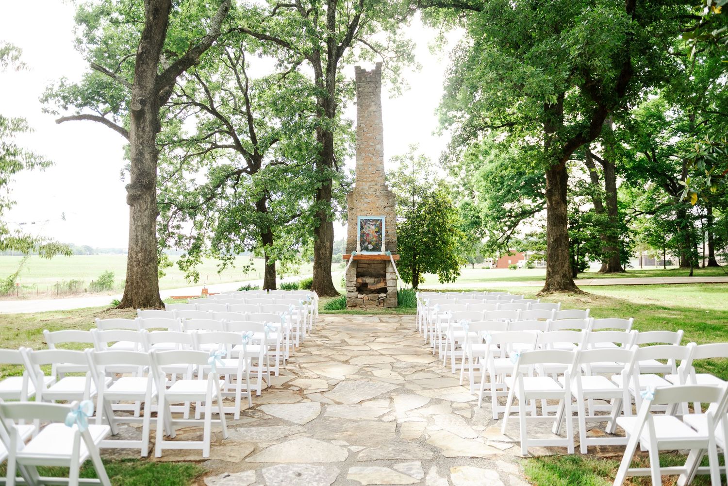 Elegant outdoor wedding ceremony setup with white chairs and stone fireplace altar under large trees.