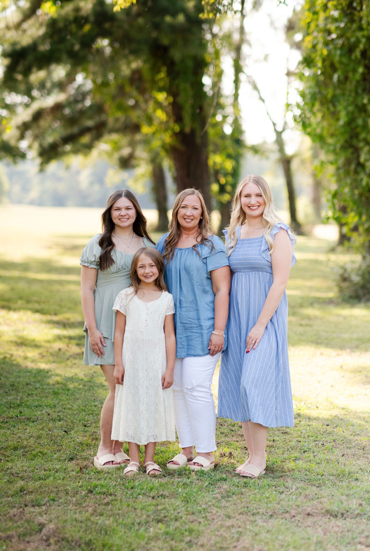 A mother poses with her three daughters in pastel dresses under green trees in a sunny park.