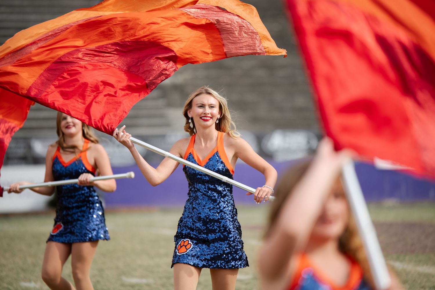 Color guard members in blue sequined costumes wave large orange flags on stadium field.