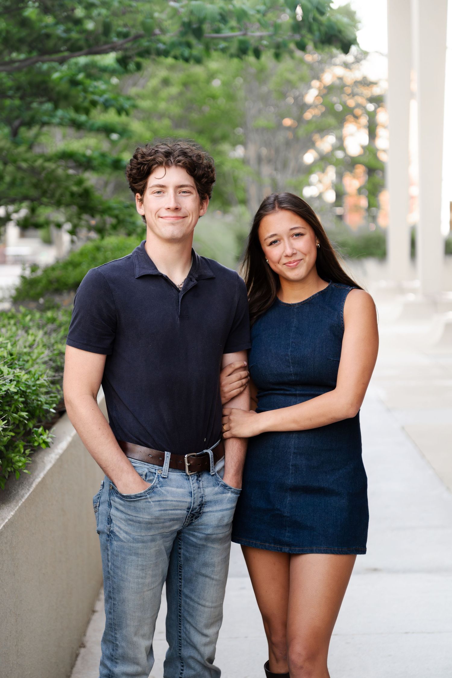 Smiling young couple posing on a sunny urban sidewalk, man in navy polo and woman in blue mini dress.