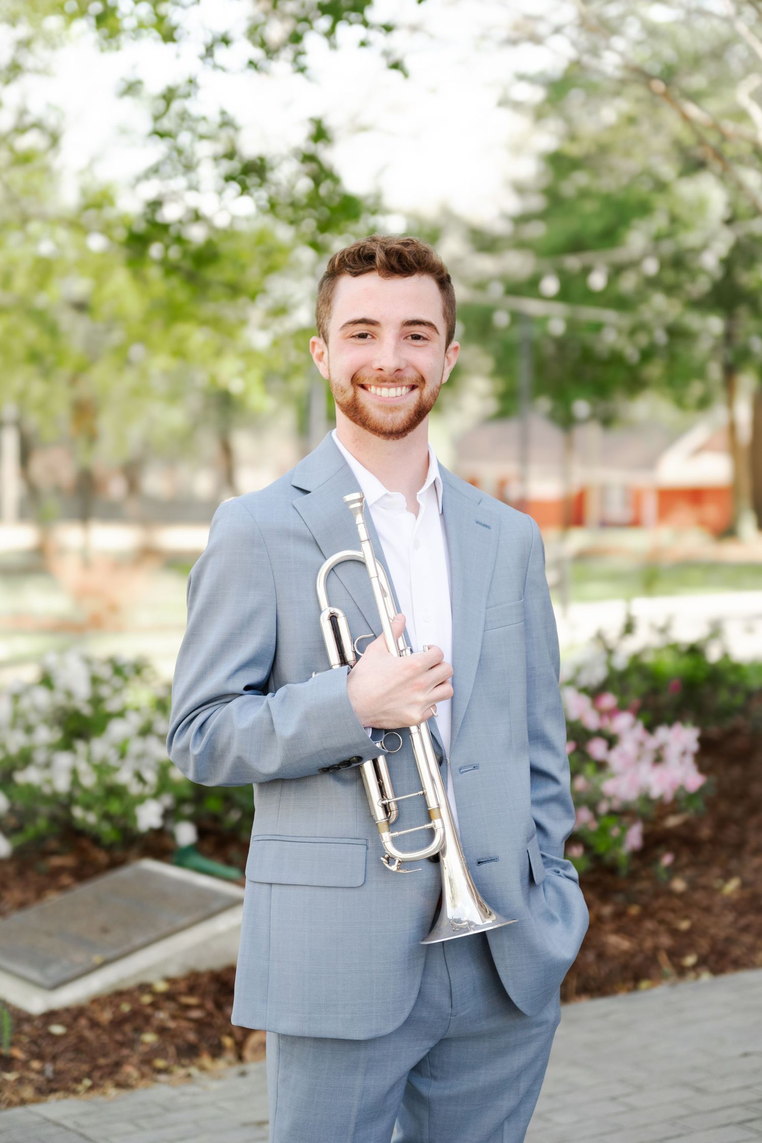 Young man in light blue suit smiling outdoors while holding a silver trumpet among garden flowers.