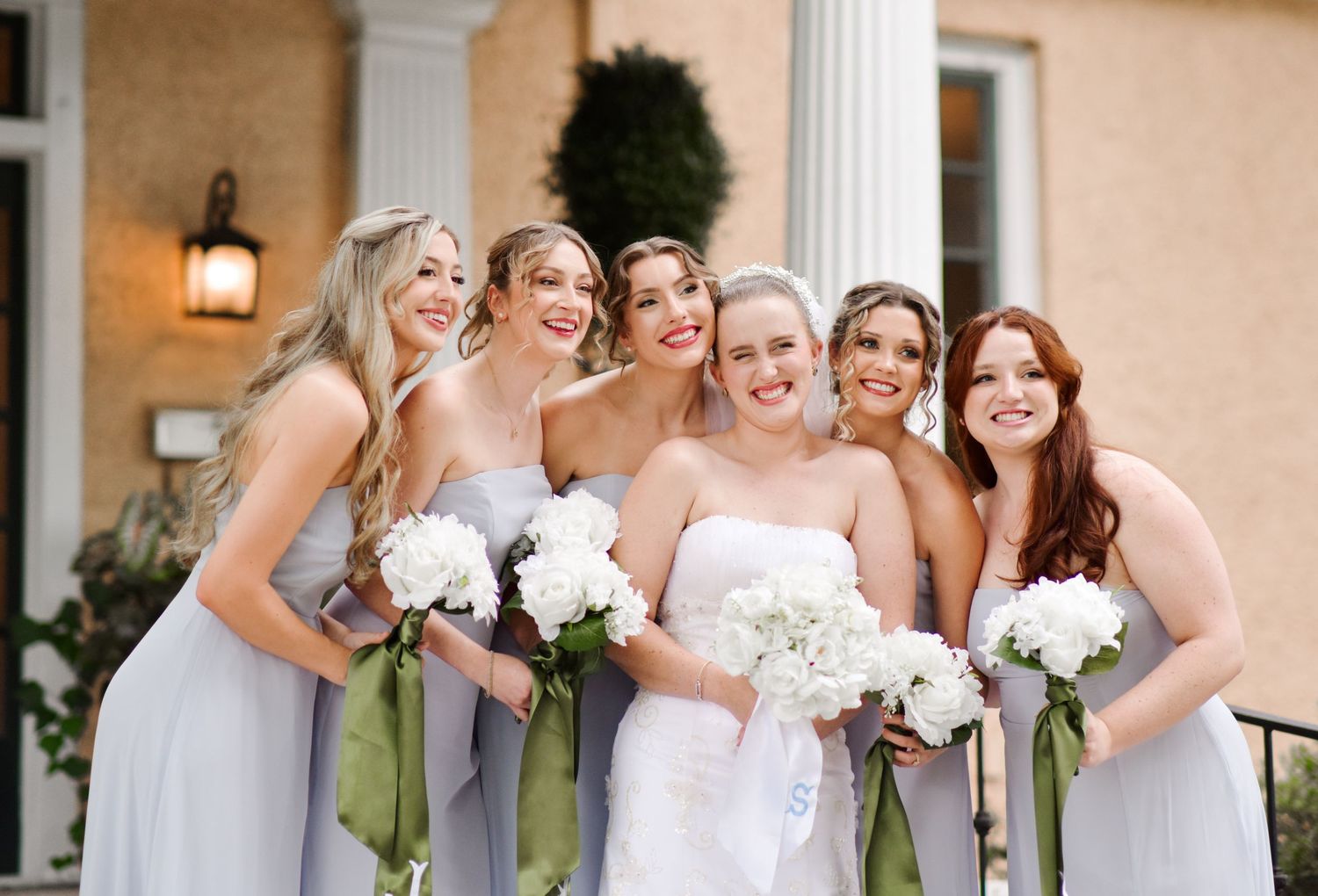 Bride with four bridesmaids in light gray dresses holding white peony bouquets outdoors.