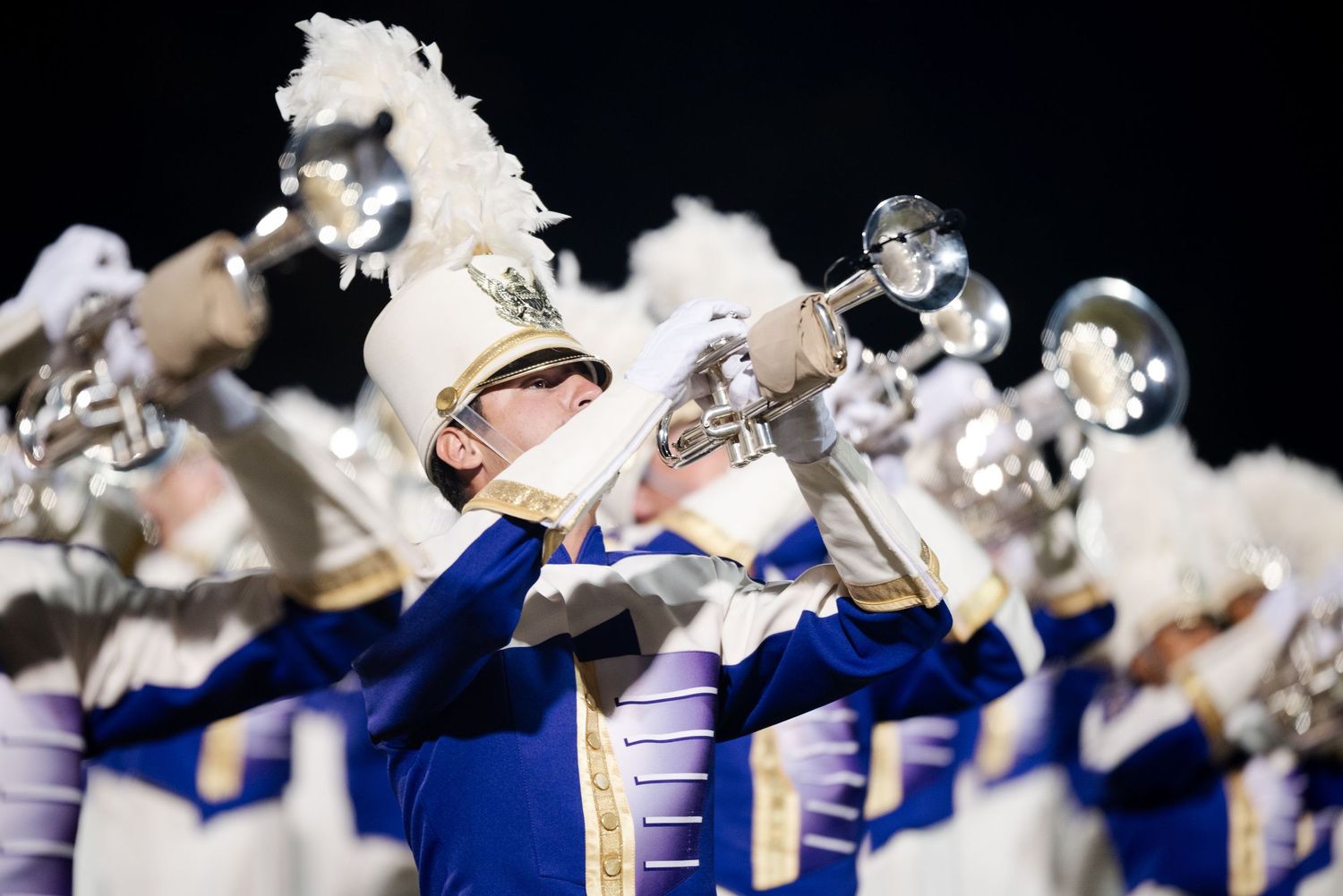 Marching band members in blue and white uniforms playing brass instruments at night.
