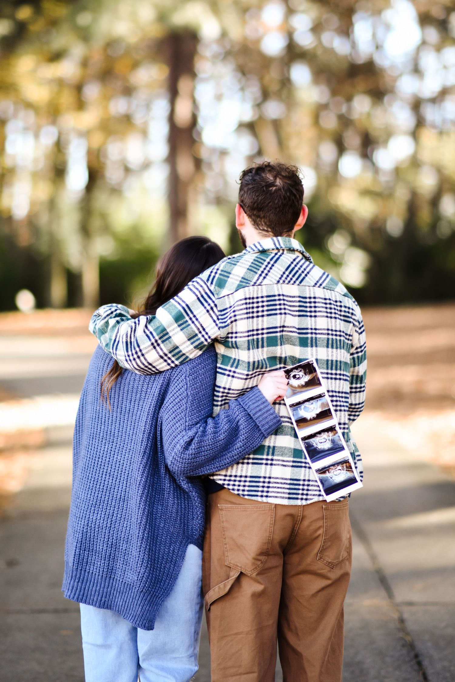 Couple seen from behind in a pine forest, man holding ultrasound photos, announcing pregnancy in a touching outdoor moment.