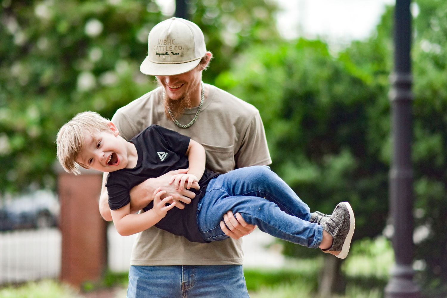 A joyful father in a tan cap cradles his giggling young son in his arms in a park.