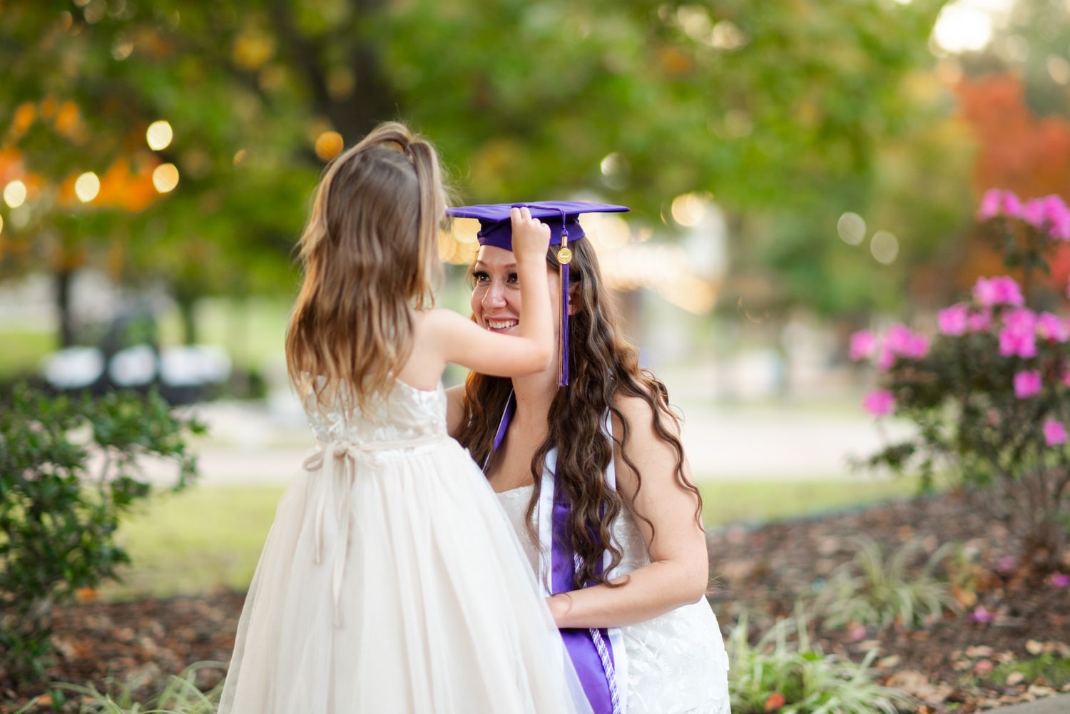 A graduate in cap and gown shares a tender moment with a young girl in a white dress outdoors.