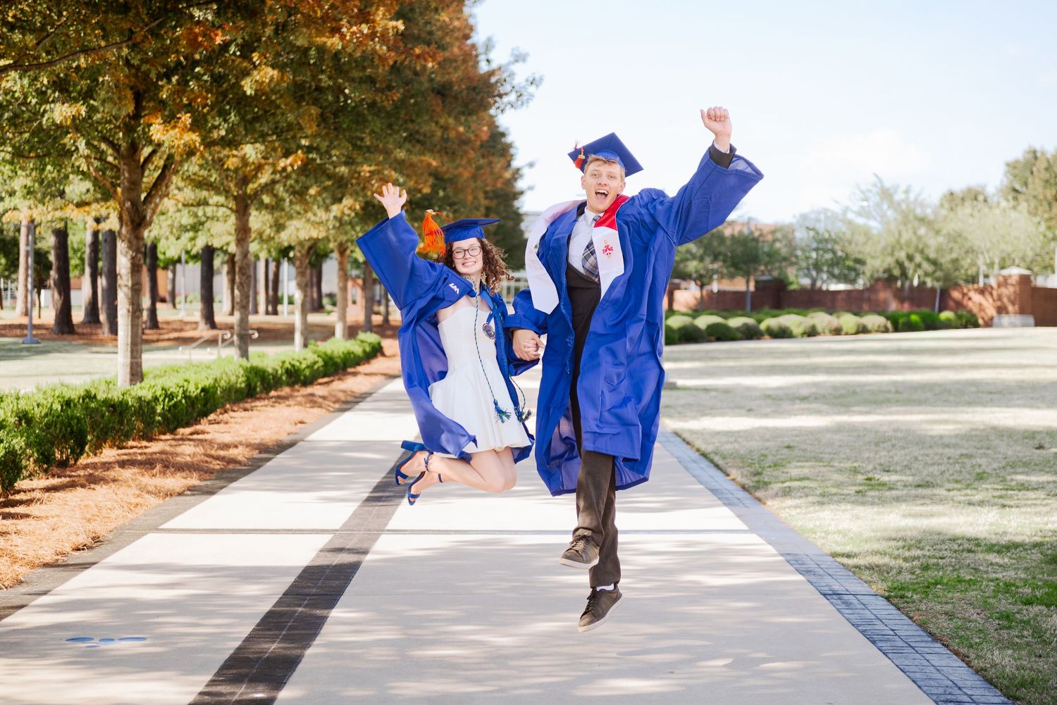 Two graduates in blue caps and gowns jumping joyfully on a tree-lined campus walkway.