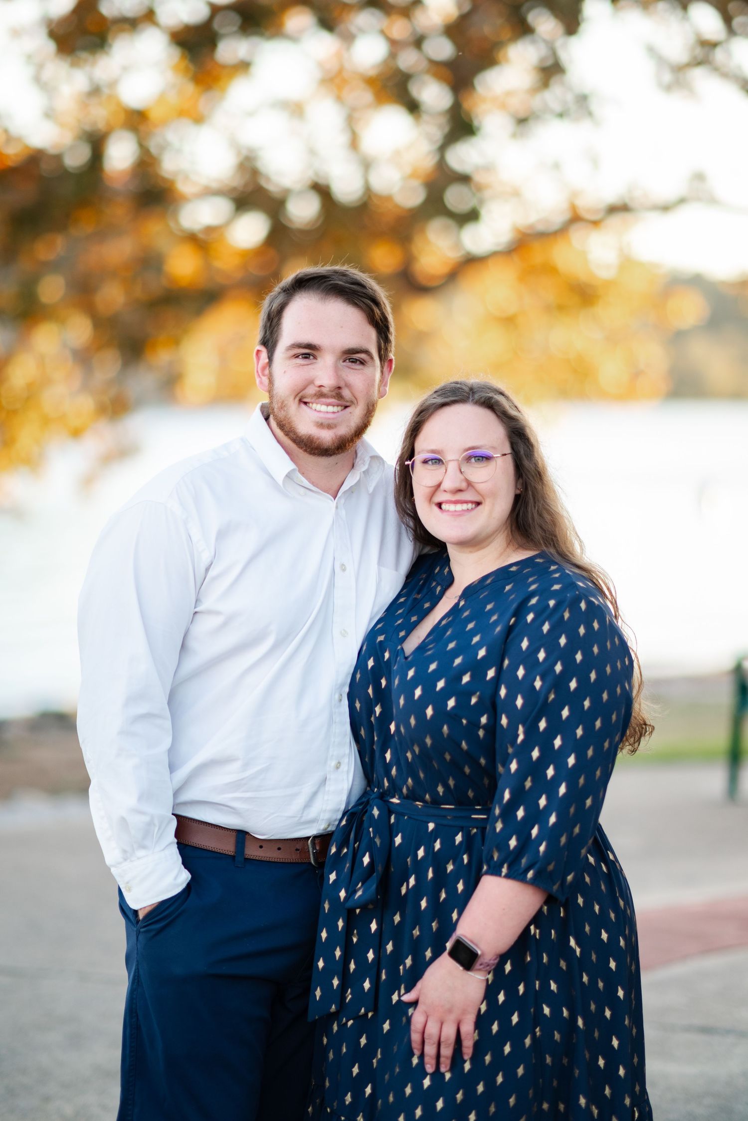 A smiling couple poses together near a waterfront with golden autumn foliage in the background.