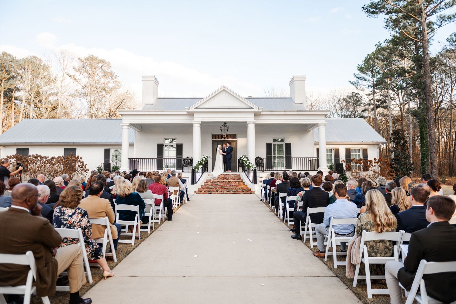 Guests seated at a sunlit outdoor wedding ceremony at a classic white colonial-style venue with tall trees.