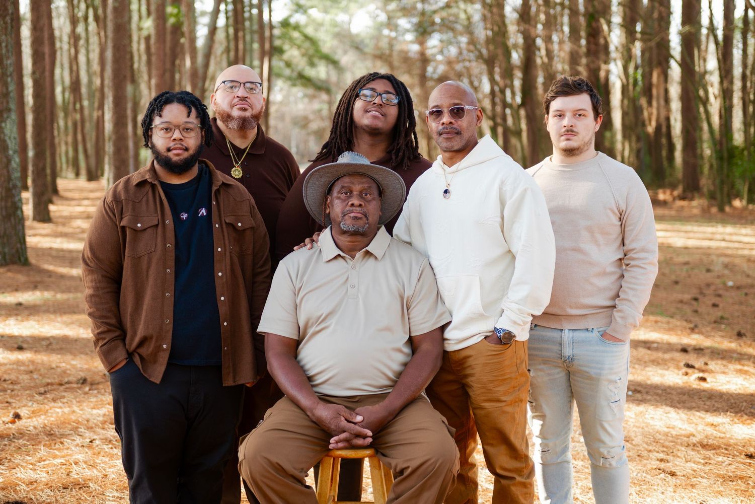 Group of six men pose seriously for portrait in pine forest with one man seated centrally.