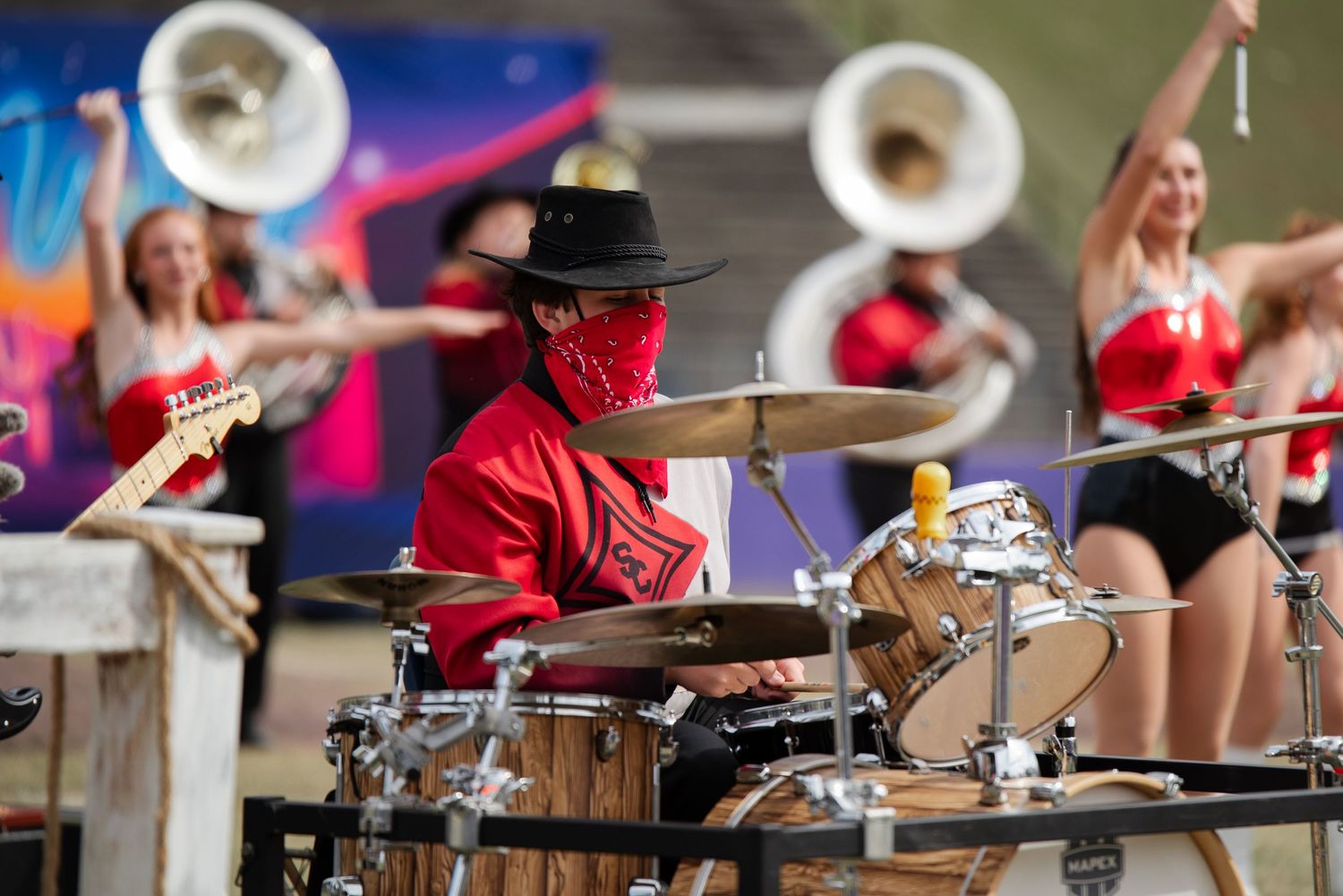 Masked drummer in red outfit performs at colorful outdoor band event with dancers.