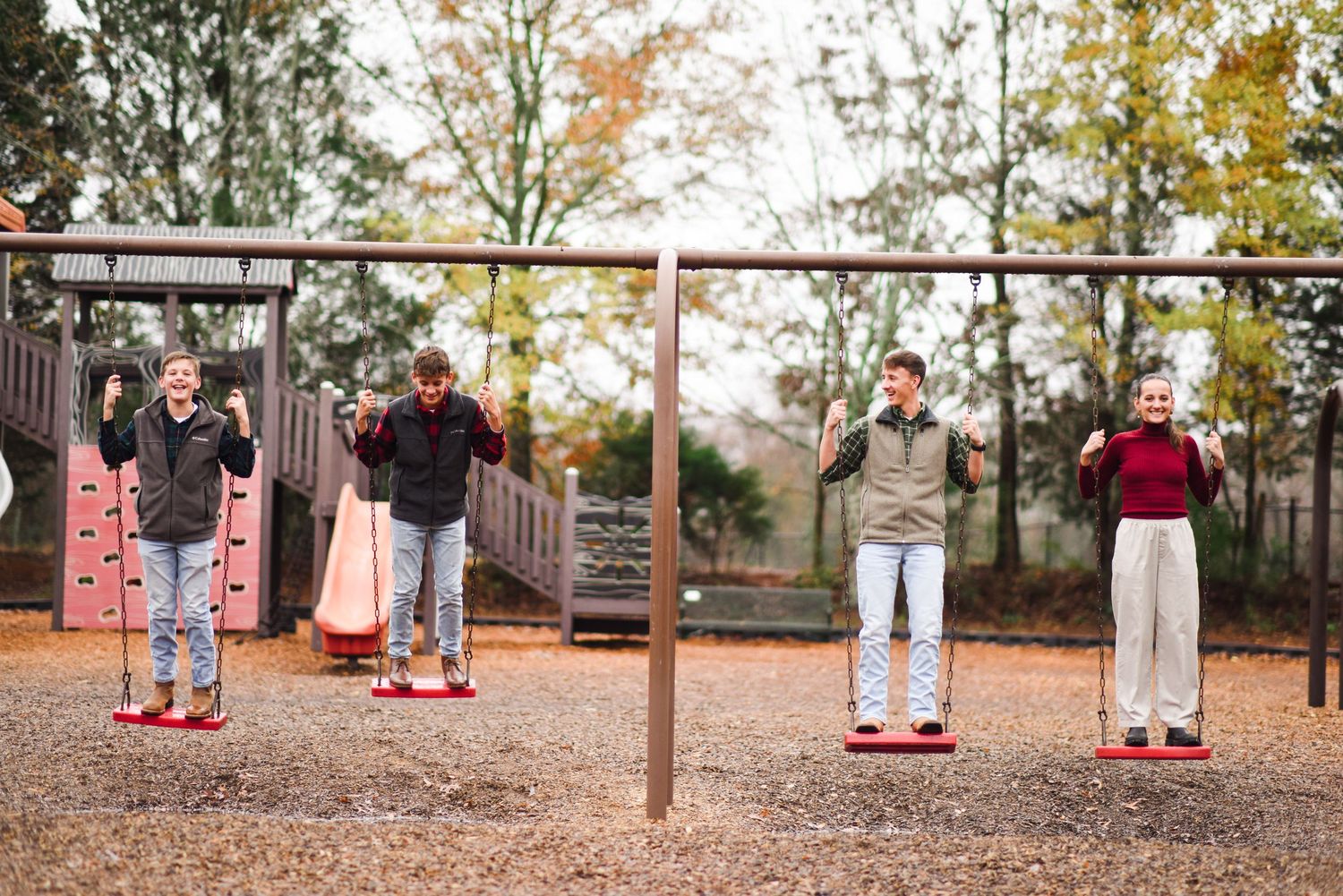 Four people joyfully swinging on playground swings surrounded by autumn foliage.