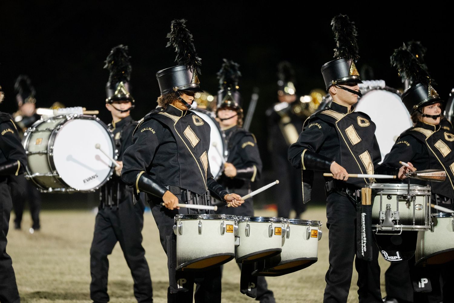 A black-uniformed marching band percussion section plays drums during a nighttime field performance.