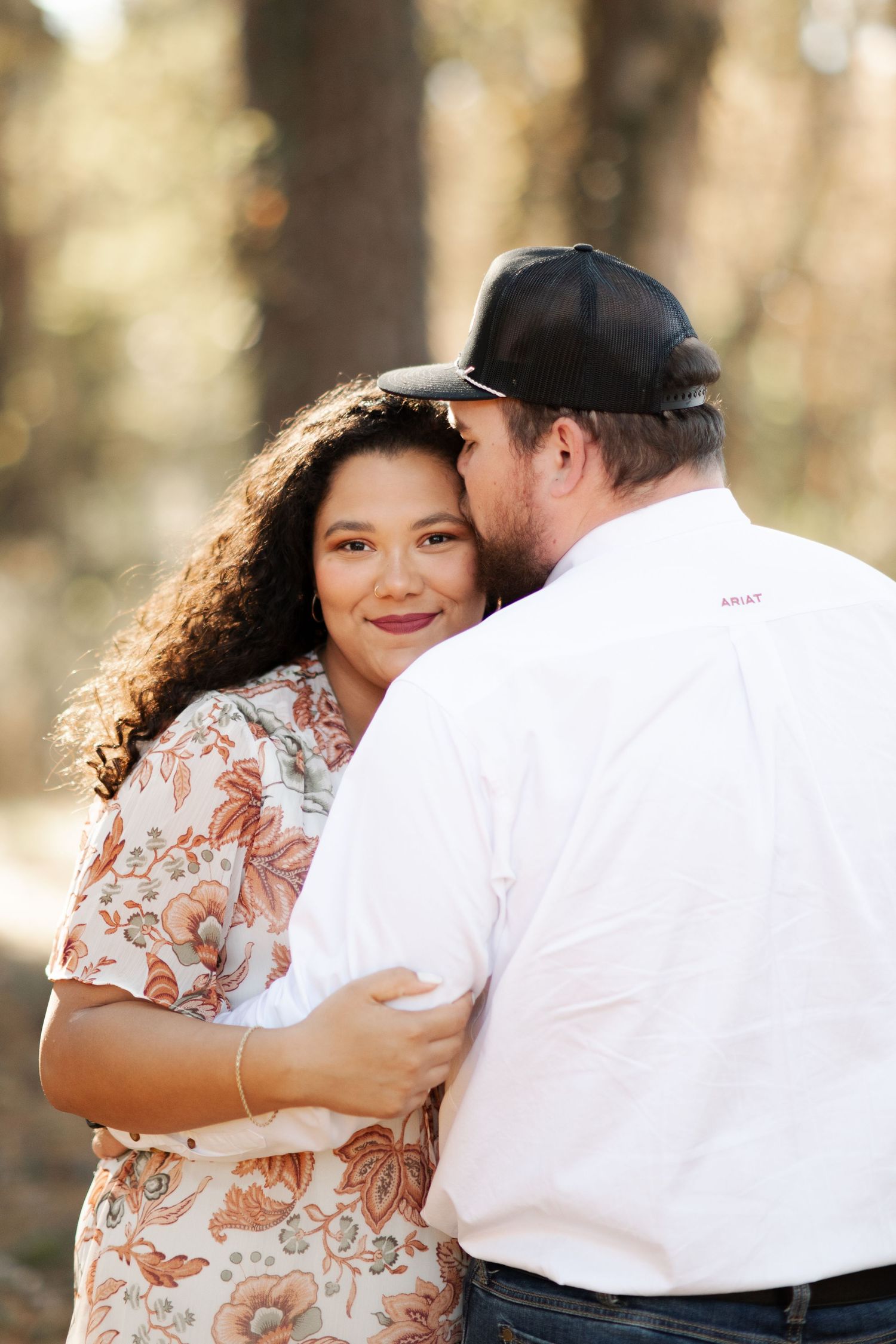 A couple embraces outdoors among pine trees, with the woman smiling at the camera.
