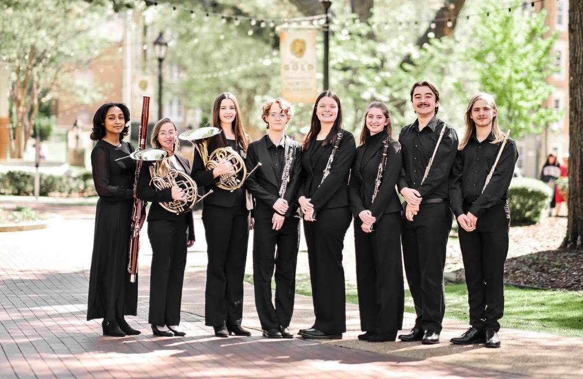 Group of eight musicians in black formal attire posing outdoors on a brick walkway.