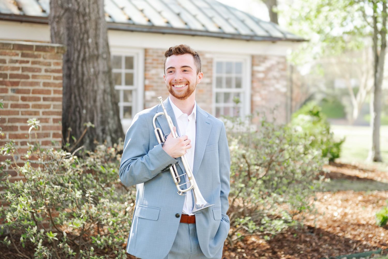 Young man in light blue suit smiling outdoors, holding a silver trumpet near a brick building.