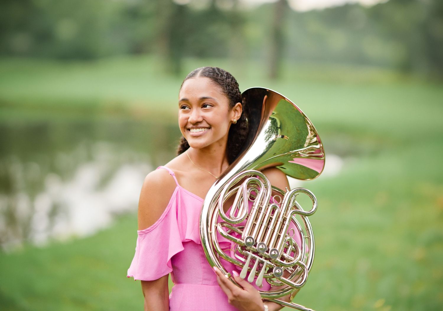 A smiling young woman in a pink dress proudly holds a French horn in a green outdoor setting.