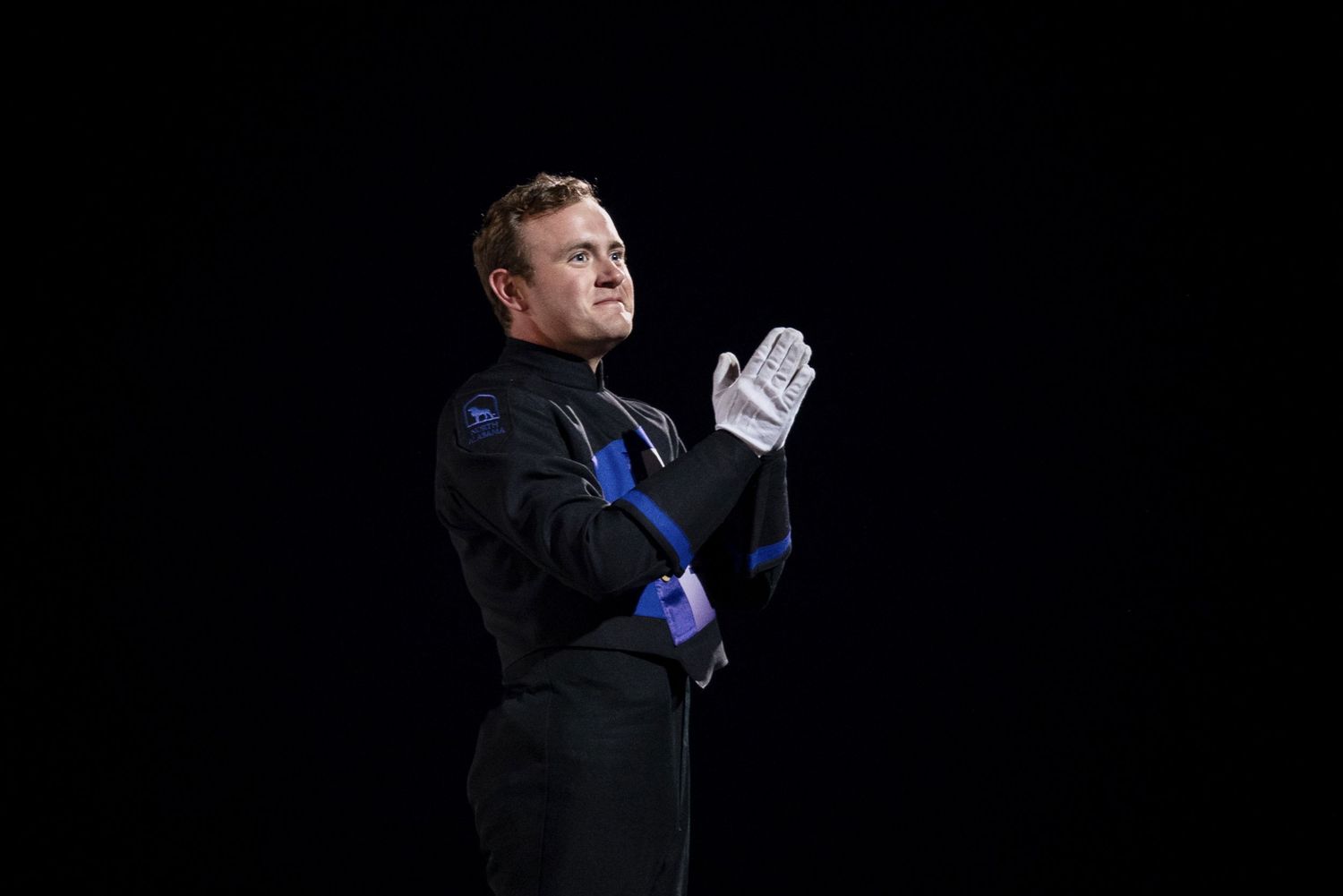 Marching band drum major in black uniform with white gloves performing against dark background.