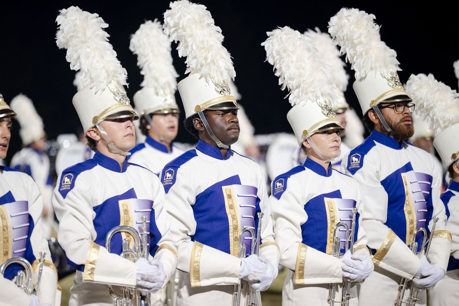 Marching band members in blue and white uniforms with tall white feathered hats standing at attention.