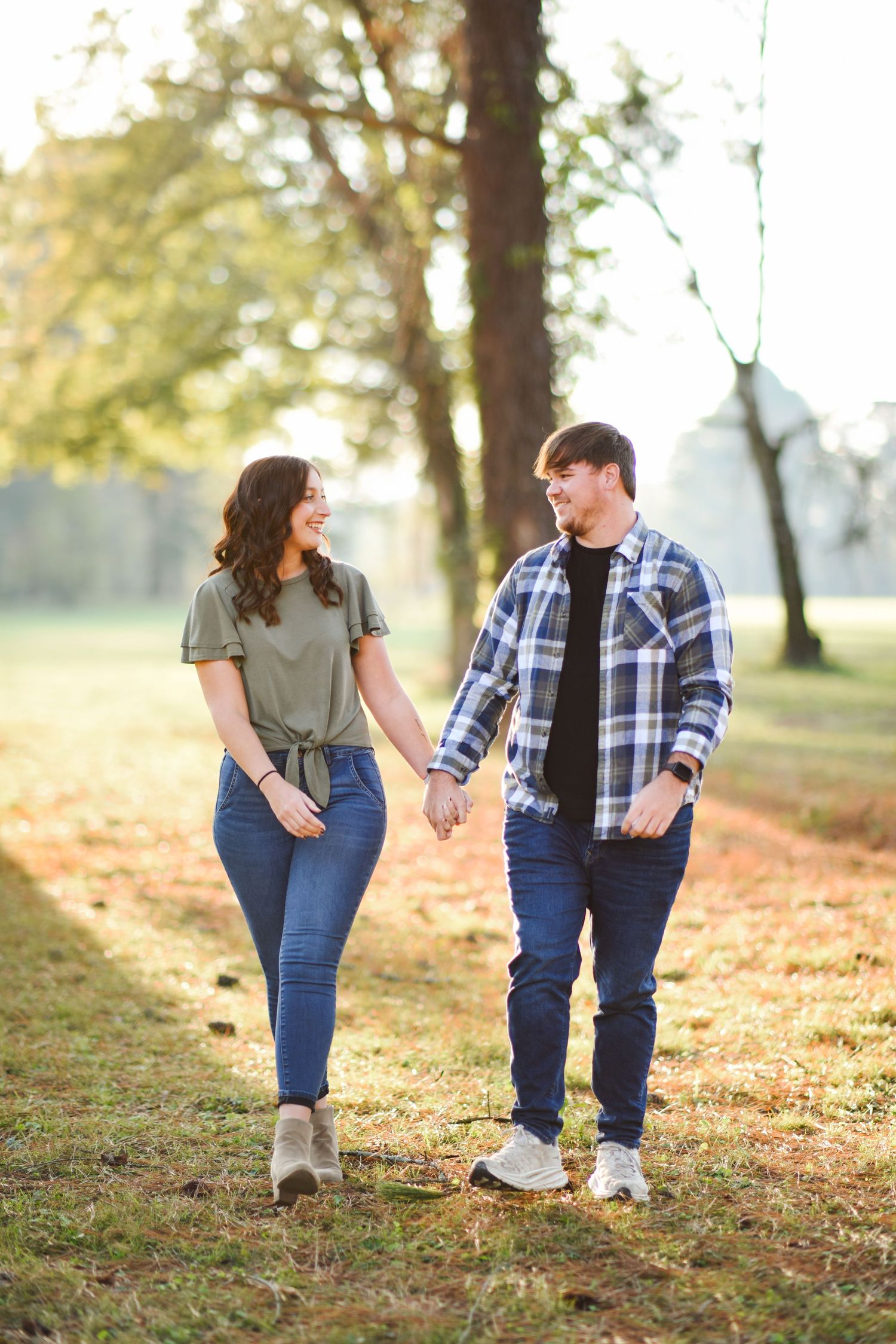 Couple holding hands and smiling at each other while walking through a golden sunlit park.