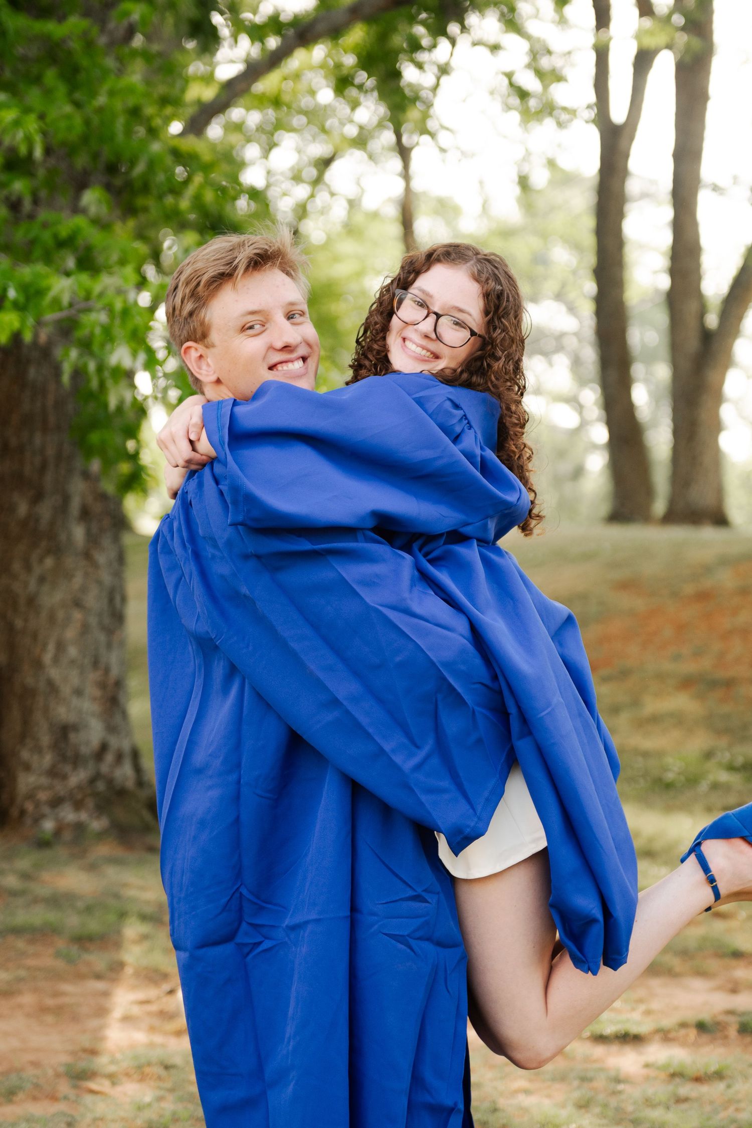 Young man in blue graduation gown carrying smiling woman with curly hair and glasses outdoors.