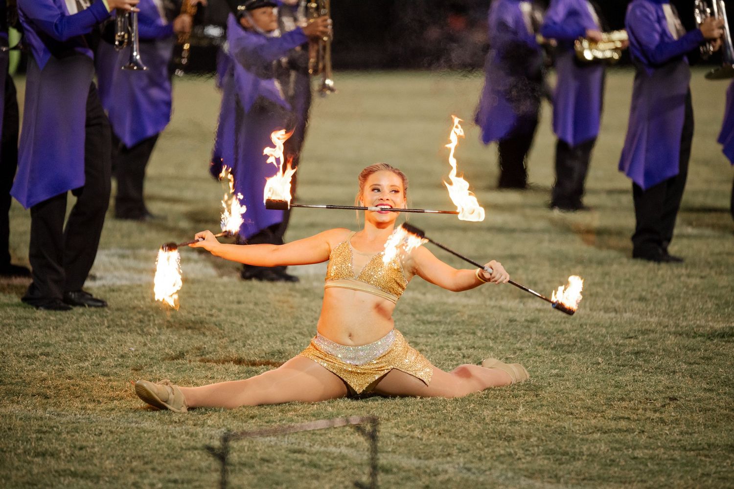 Female fire twirler performing splits with flaming batons at marching band show.