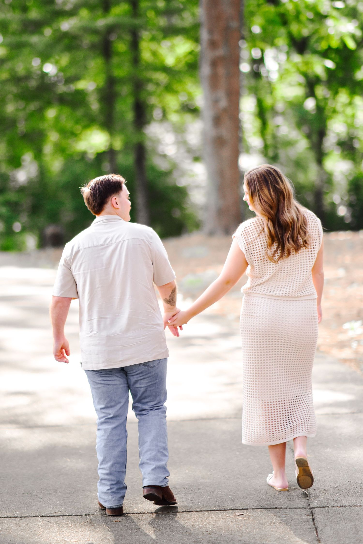 Couple holding hands walking away on a path surrounded by lush green trees in a park.