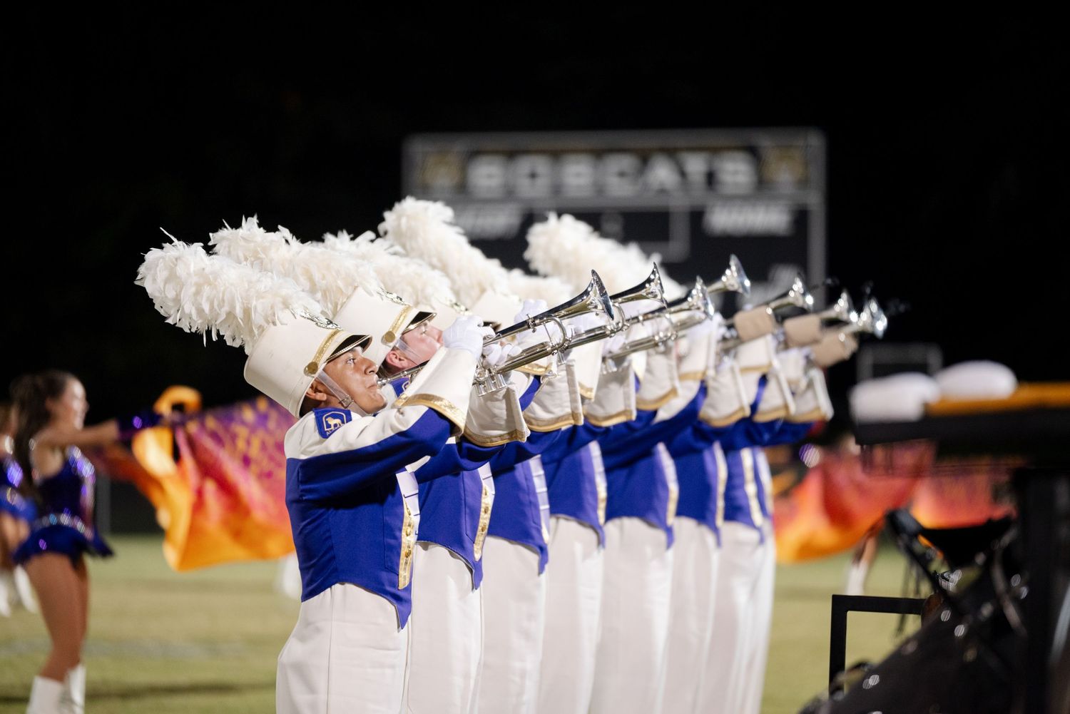 Marching band members in blue and white uniforms playing trumpets on a football field at night.