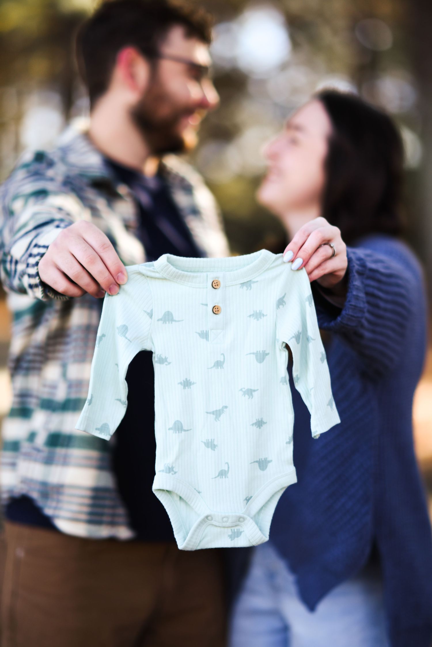 Smiling couple holding up a light blue dinosaur print baby onesie outdoors.